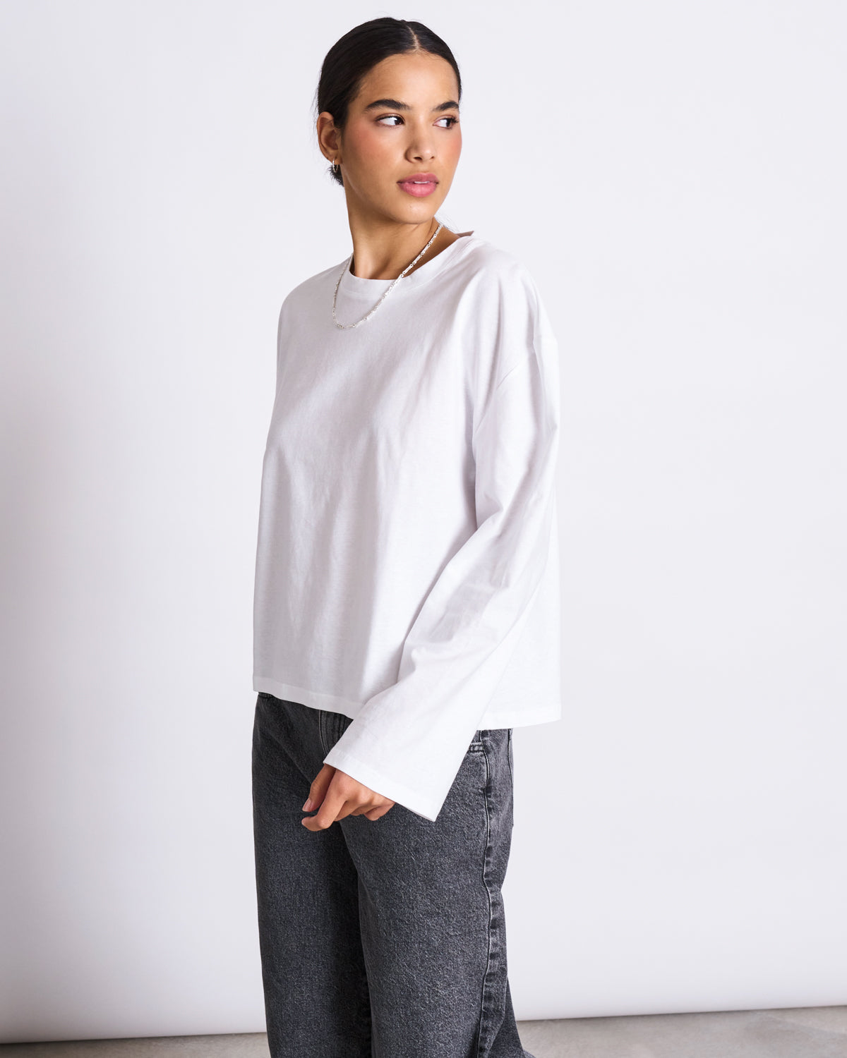 A person with dark hair tied back wears an oversized BOXY LONGSLEEVE ANKIRA WHITE by jan-n-june in organic cotton with faded black jeans, standing against a white background and looking to the side.