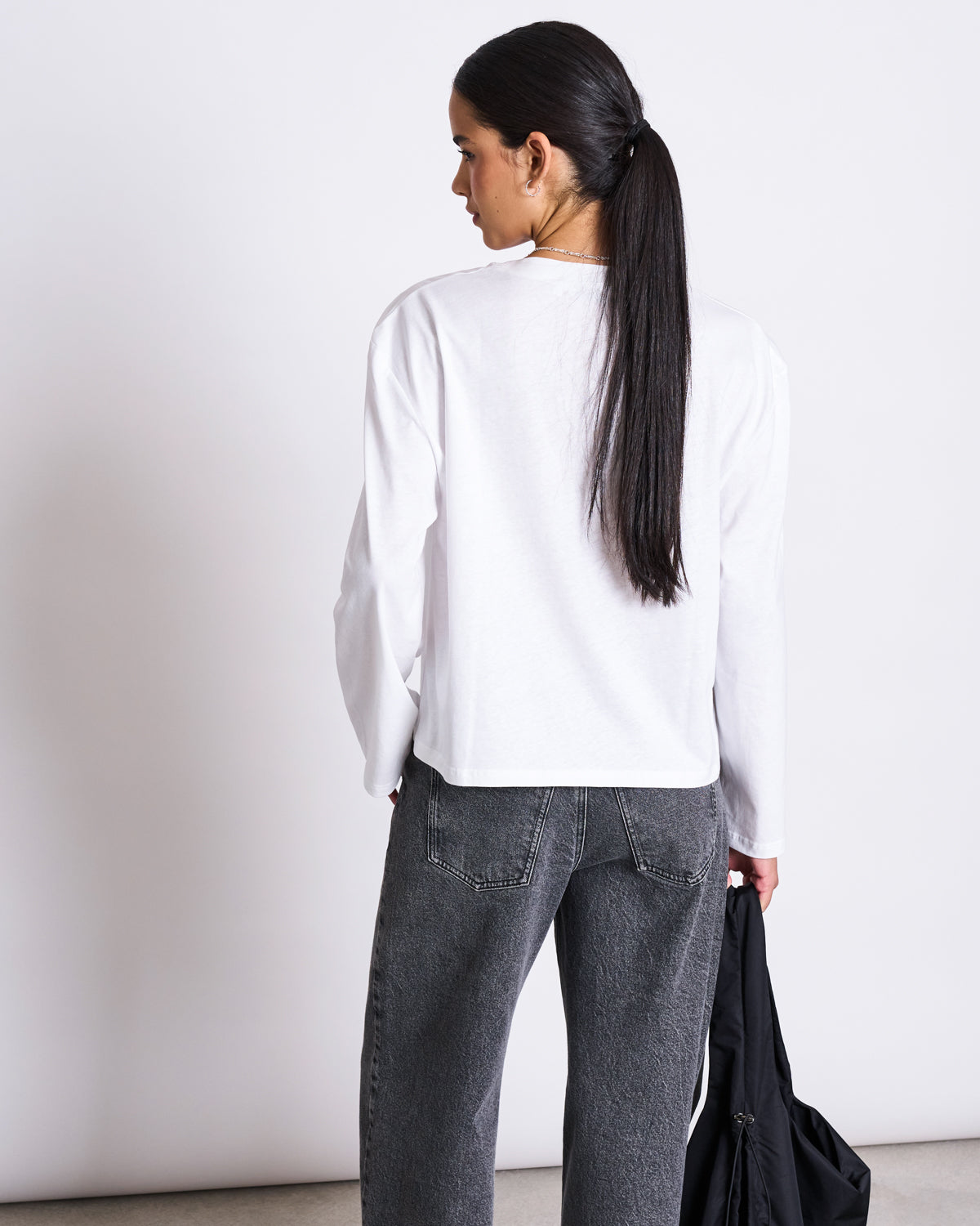 A woman with long dark hair in a low ponytail wears gray jeans and the jan-n-june BOXY LONGSLEEVE ANKIRA WHITE shirt, standing with her back to the camera and holding a black jacket in her left hand against a plain white background.