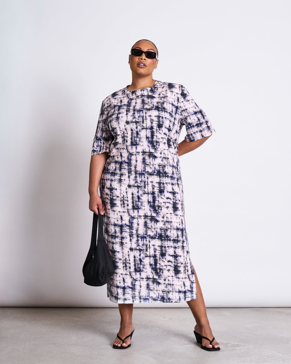 A person stands confidently against a plain white background wearing jan-n-junes SHIRT DRESS CROVIE MELTED PRINT GOTS, styled with sunglasses, a black bag, and black sandals.
