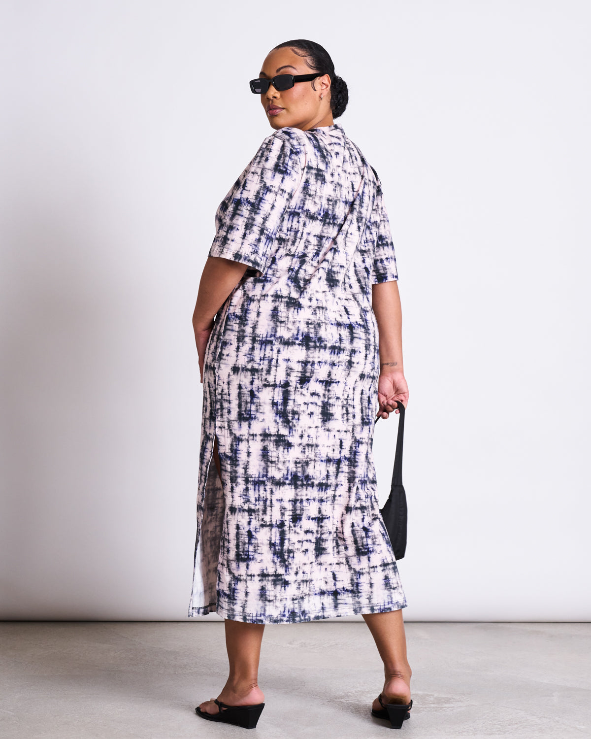 A woman in sunglasses wears the jan-n-june SHIRT DRESS CROVIE MELTED PRINT GOTS—an oversized, organic cotton midi dress. She stands sideways, looking over her shoulder, with a black handbag and heeled sandals against a white background.