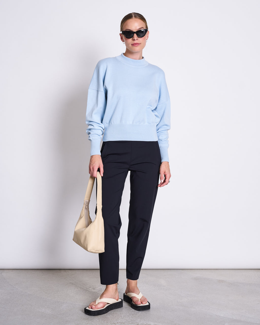 A woman in jan-n-june PANTS STRAIGHT BLACK, a light blue sweater, black metallic-strap sandals, sunglasses, and a beige handbag stands against a white background.