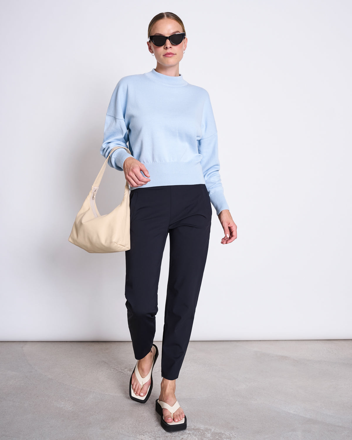 A woman wearing sunglasses, a light blue sweater, white sandals, and carrying a beige shoulder bag confidently walks against a plain white background in jan-n-june’s PANTS STRAIGHT BLACK.
