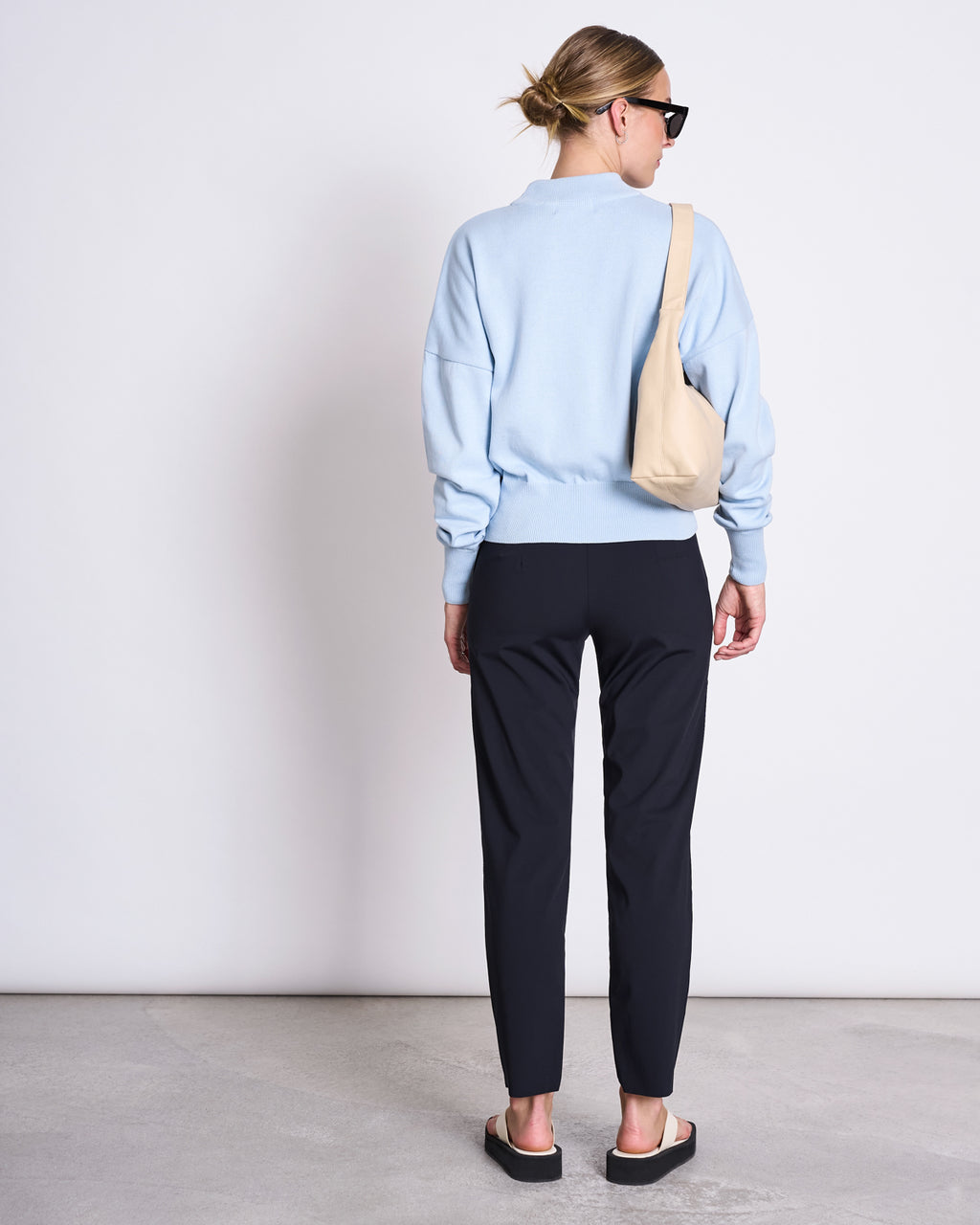 A woman stands facing away in jan-n-junes PANTS STRAIGHT BLACK, paired with a light blue sweater, black slide sandals, sunglasses, a beige shoulder bag, and her hair in a bun against a plain light background.