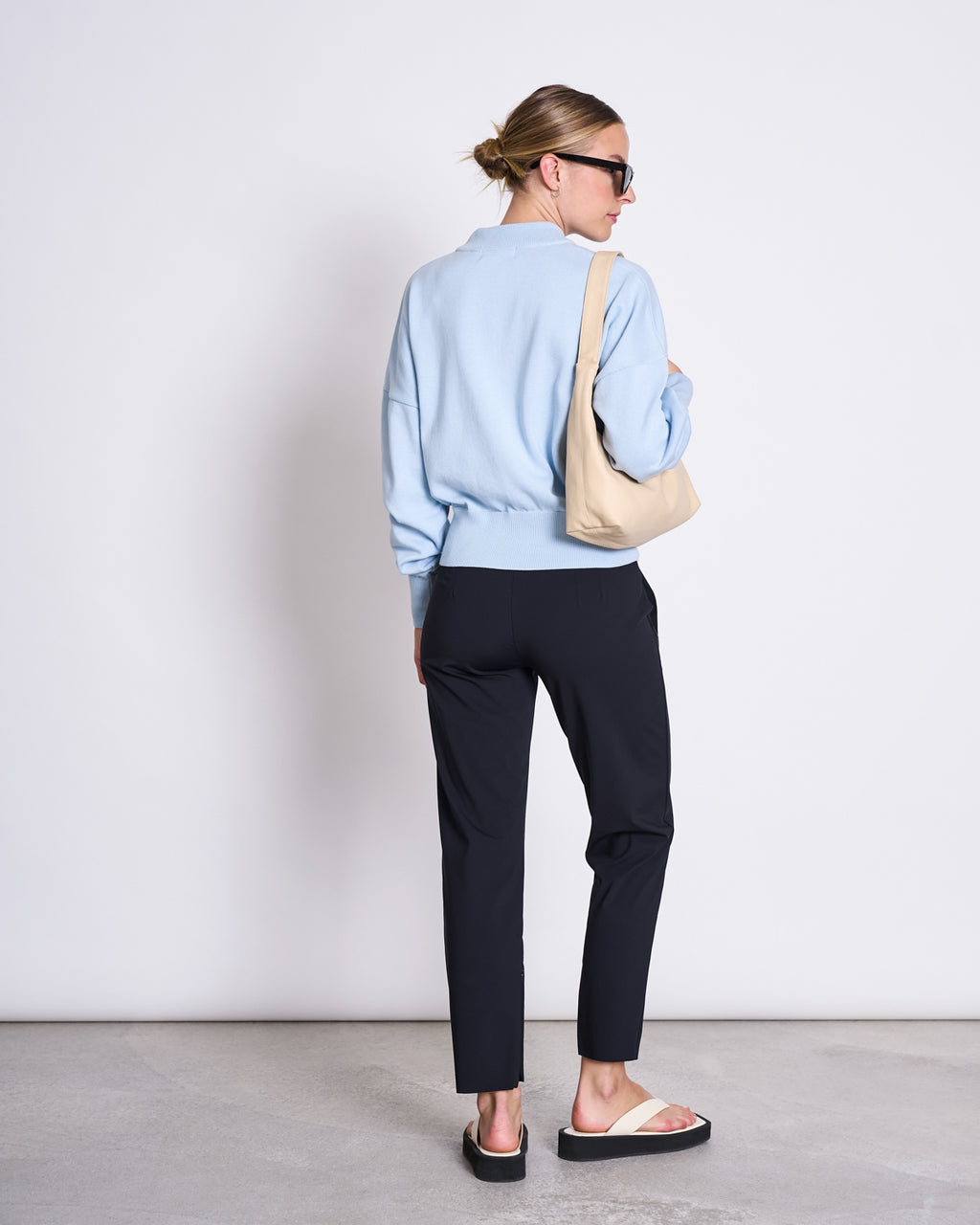 A woman with blonde hair in a bun, black sunglasses, light blue sweater, and sandals faces away against a plain background, carrying a beige bag and wearing jan-n-june PANTS STRAIGHT BLACK.