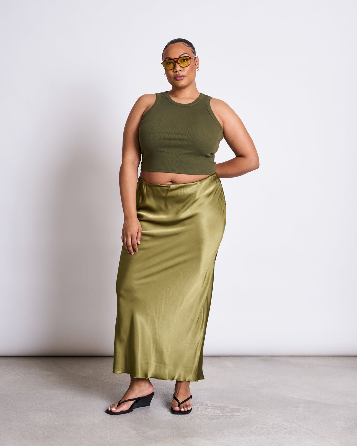 A woman in yellow sunglasses, a jan-n-june ORGANIC COTTON TANK TOP PORTO DARK OLIVE GOTS, and a shiny olive green satin skirt stands on a concrete floor against a plain white background.