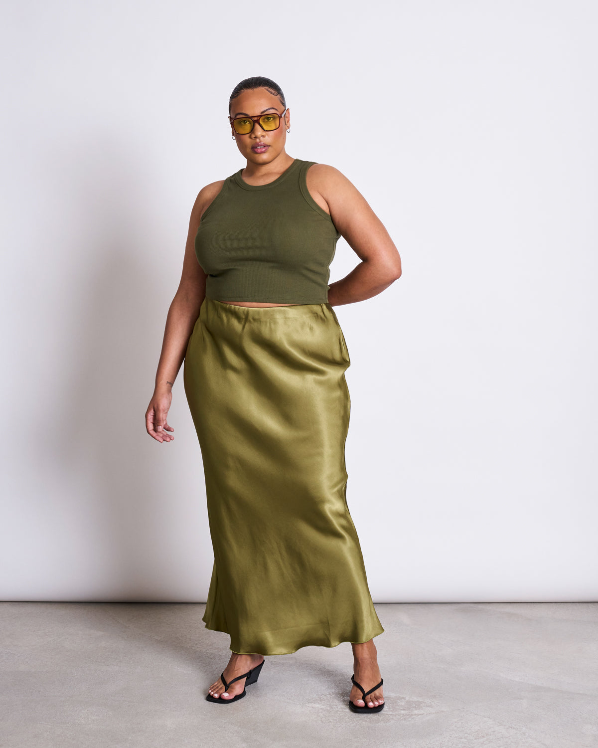 A person poses confidently in black heeled sandals, yellow sunglasses, a shiny olive green maxi skirt, and the jan-n-june ORGANIC COTTON TANK TOP PORTO DARK OLIVE GOTS, standing against a plain white background.
