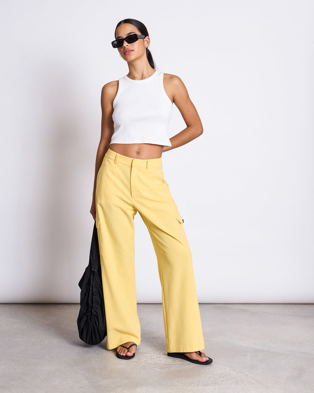 A woman poses against a plain backdrop wearing jan-n-junes CARGO PANTS FANO BUTTERCUP, a pair of high-waisted yellow wide-leg pants, styled with a white crop top, black sandals, sunglasses, and a large black bag.