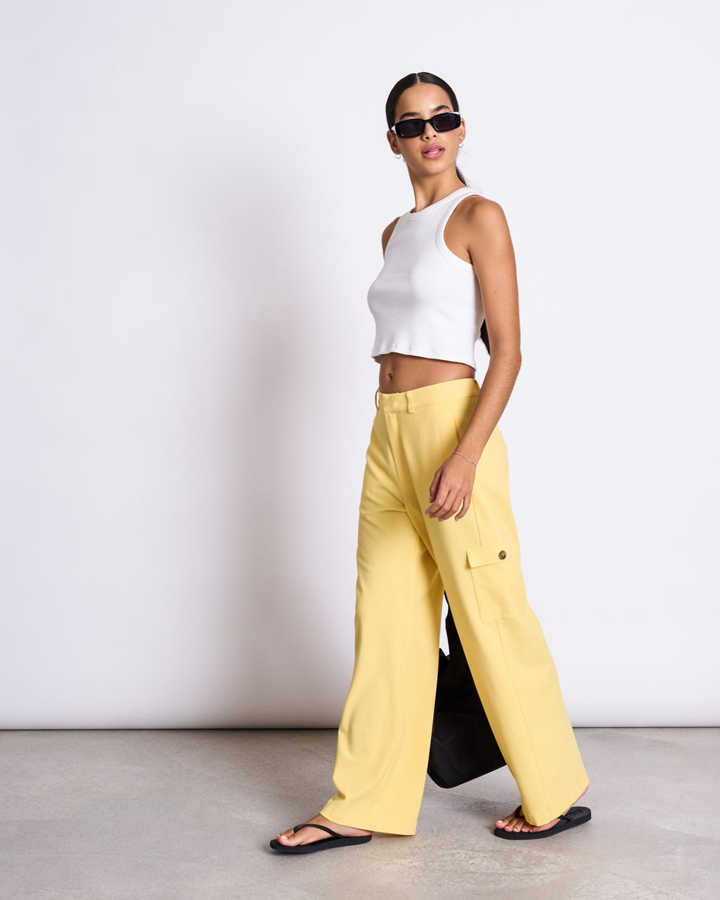 A woman in jan-n-june’s CARGO PANTS FANO BUTTERCUP, a white sleeveless crop top, black sandals, and sunglasses walks indoors against a plain white backdrop, carrying a black bag.