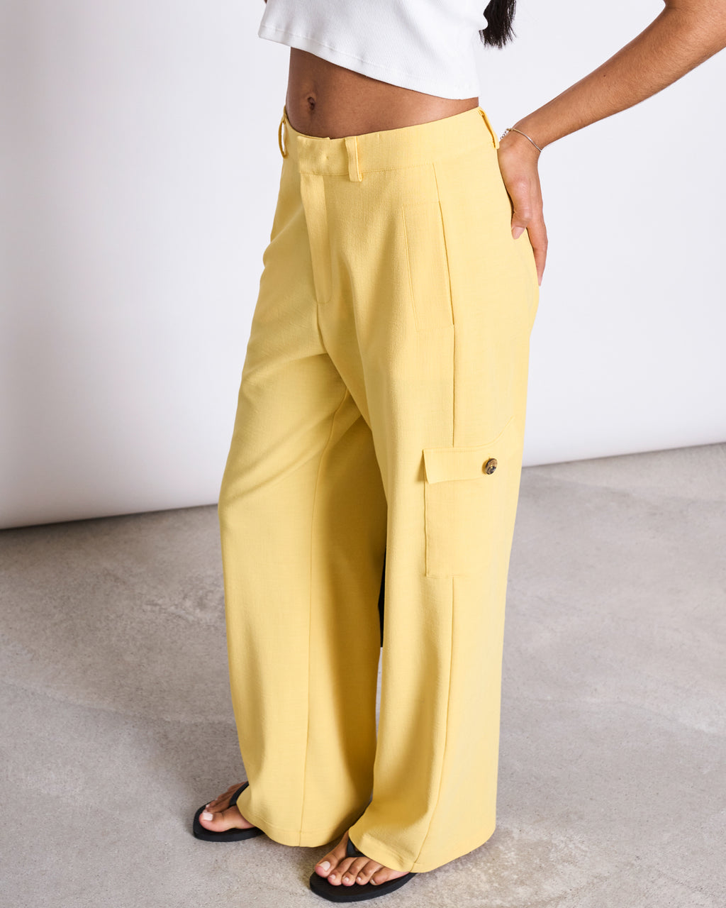 A person wears jan-n-junes CARGO PANTS FANO BUTTERCUP—high-waisted, wide-leg yellow EcoVero cargo pants with side pockets—paired with a white crop top, standing on a light gray textured floor, showing only their lower half.