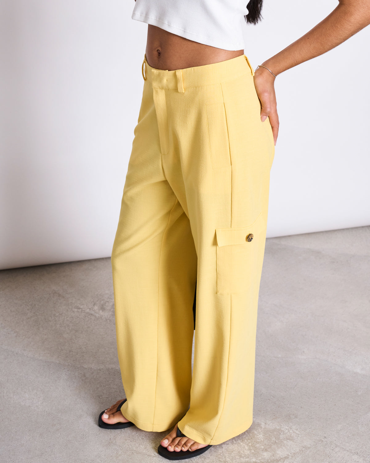 A person wears jan-n-junes CARGO PANTS FANO BUTTERCUP—high-waisted, wide-leg yellow EcoVero cargo pants with side pockets—paired with a white crop top, standing on a light gray textured floor, showing only their lower half.