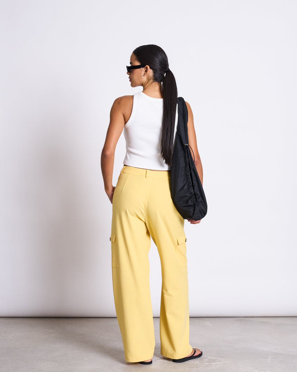 A woman with long dark hair in a ponytail wears the jan-n-june CARGO PANTS FANO BUTTERCUP, a white sleeveless top, black sandals, sunglasses, and carries a black bag over her shoulder. She stands facing away from the camera against a plain background.