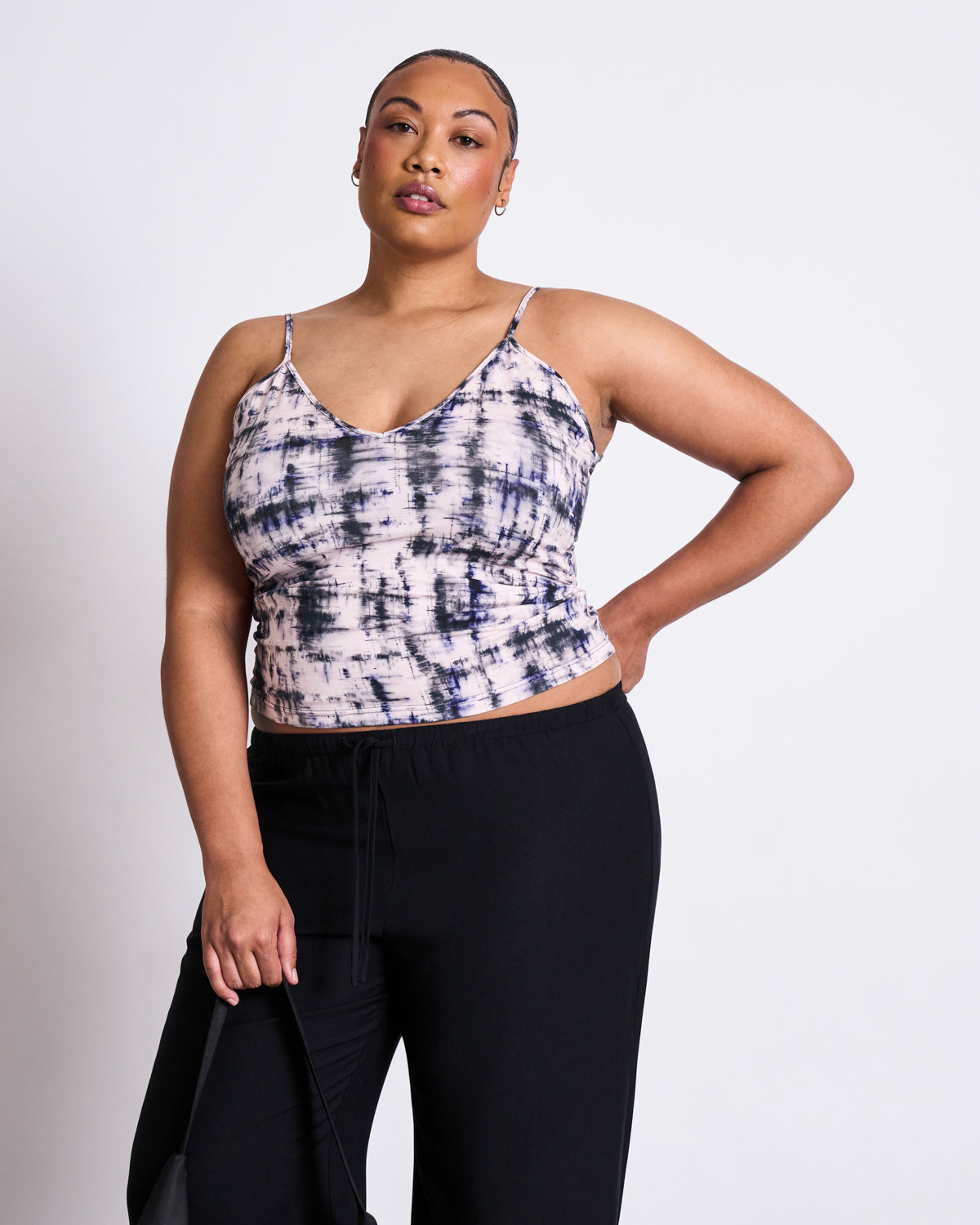 A woman poses confidently in the jan-n-june TOP MATILDA MELTED PRINT GOTS with thin straps and black pants against a plain white background, one hand on her hip as she gazes at the camera.