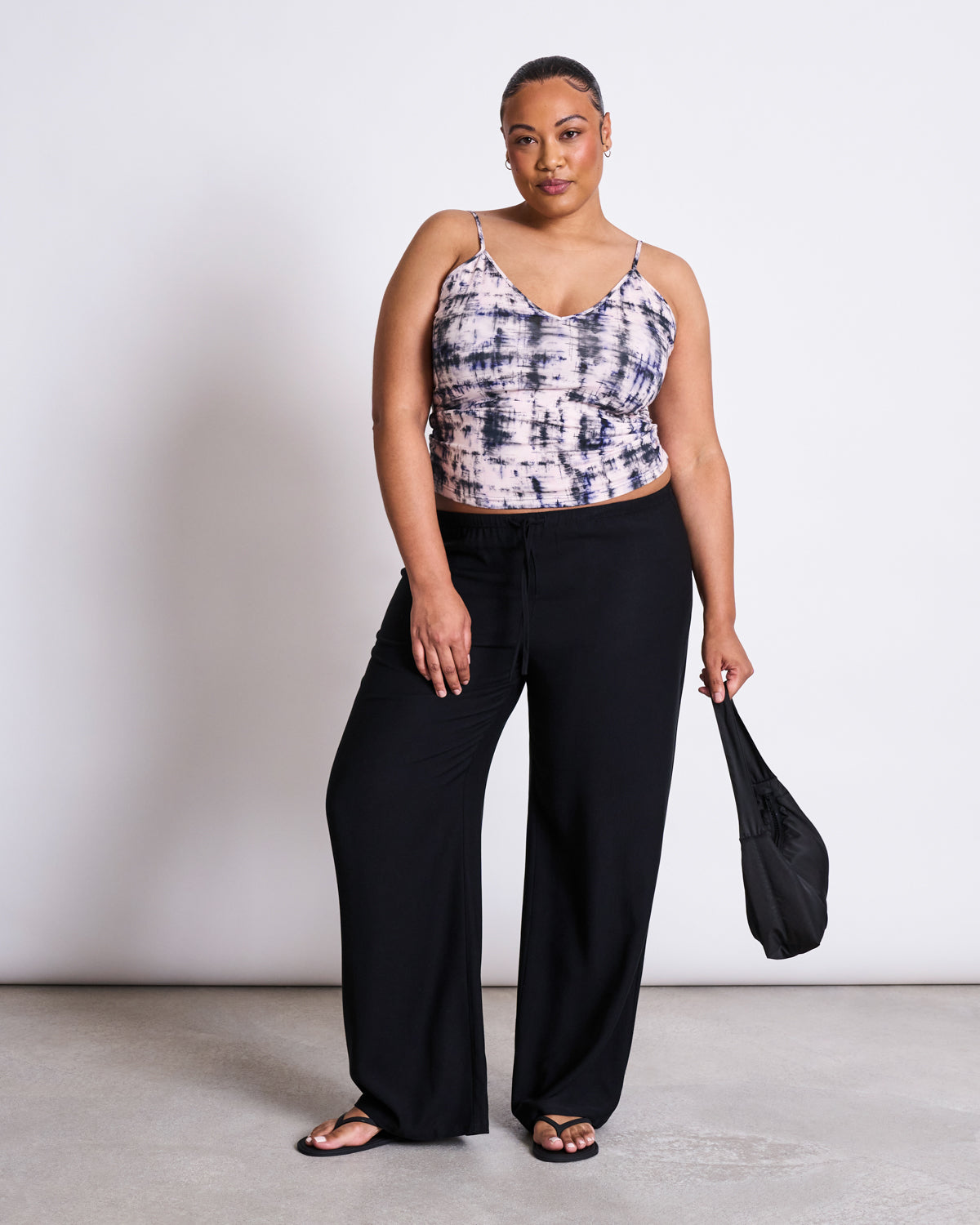 A woman stands confidently against a plain background, wearing the jan-n-june TOP MATILDA MELTED PRINT GOTS tank, loose black pants, sandals, and holding a black bag with her hair pulled back.