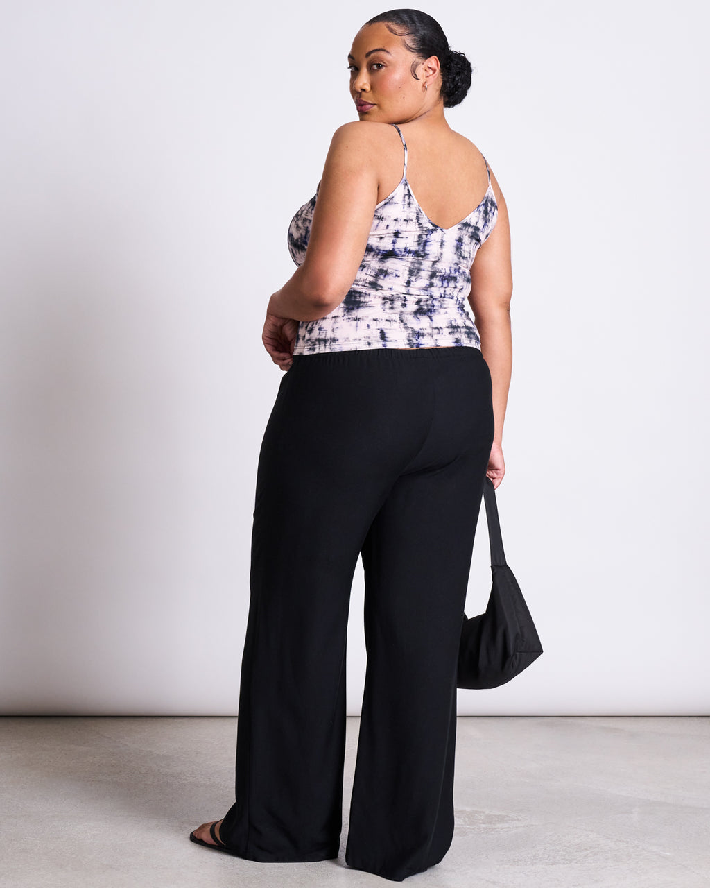 A woman with her hair in a low bun looks over her shoulder, wearing the jan-n-june TOP MATILDA MELTED PRINT GOTS, black wide-leg pants, sandals, and holding a small black bag against a plain light background.