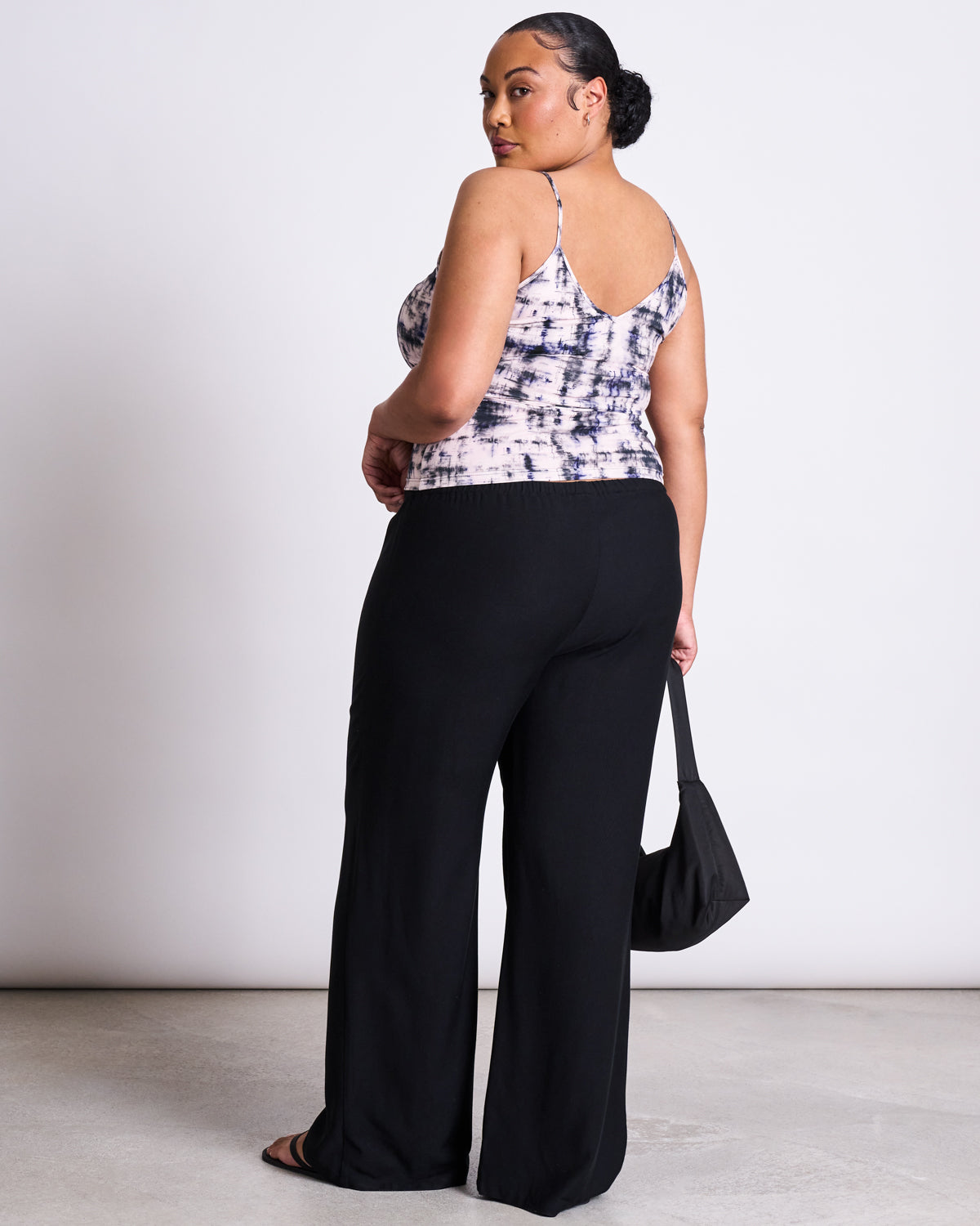 A woman with her hair in a low bun looks over her shoulder, wearing the jan-n-june TOP MATILDA MELTED PRINT GOTS, black wide-leg pants, sandals, and holding a small black bag against a plain light background.