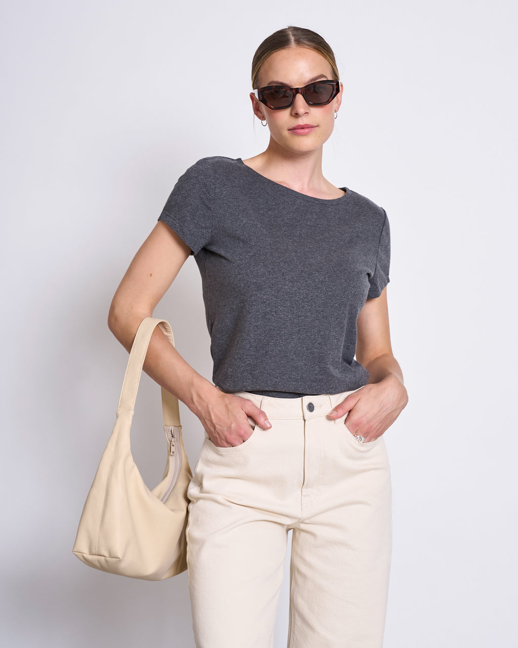 A woman stands against a plain white background, one hand in her pocket, wearing the jan-n-june T-SHIRT OTTO DARK GREY GOTS, off-white pants, dark sunglasses, and carrying a beige shoulder bag.