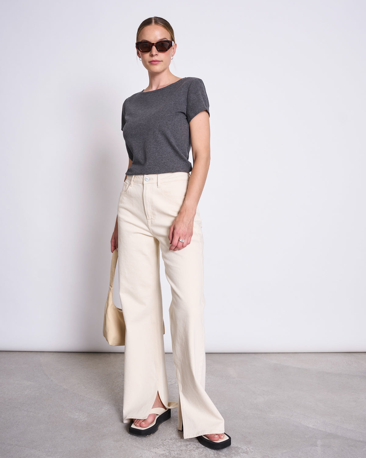 A woman in the jan-n-june T-SHIRT OTTO DARK GREY GOTS, cream wide-leg pants, black sandals and sunglasses stands against a light background, holding a beige slouchy bag at her side.