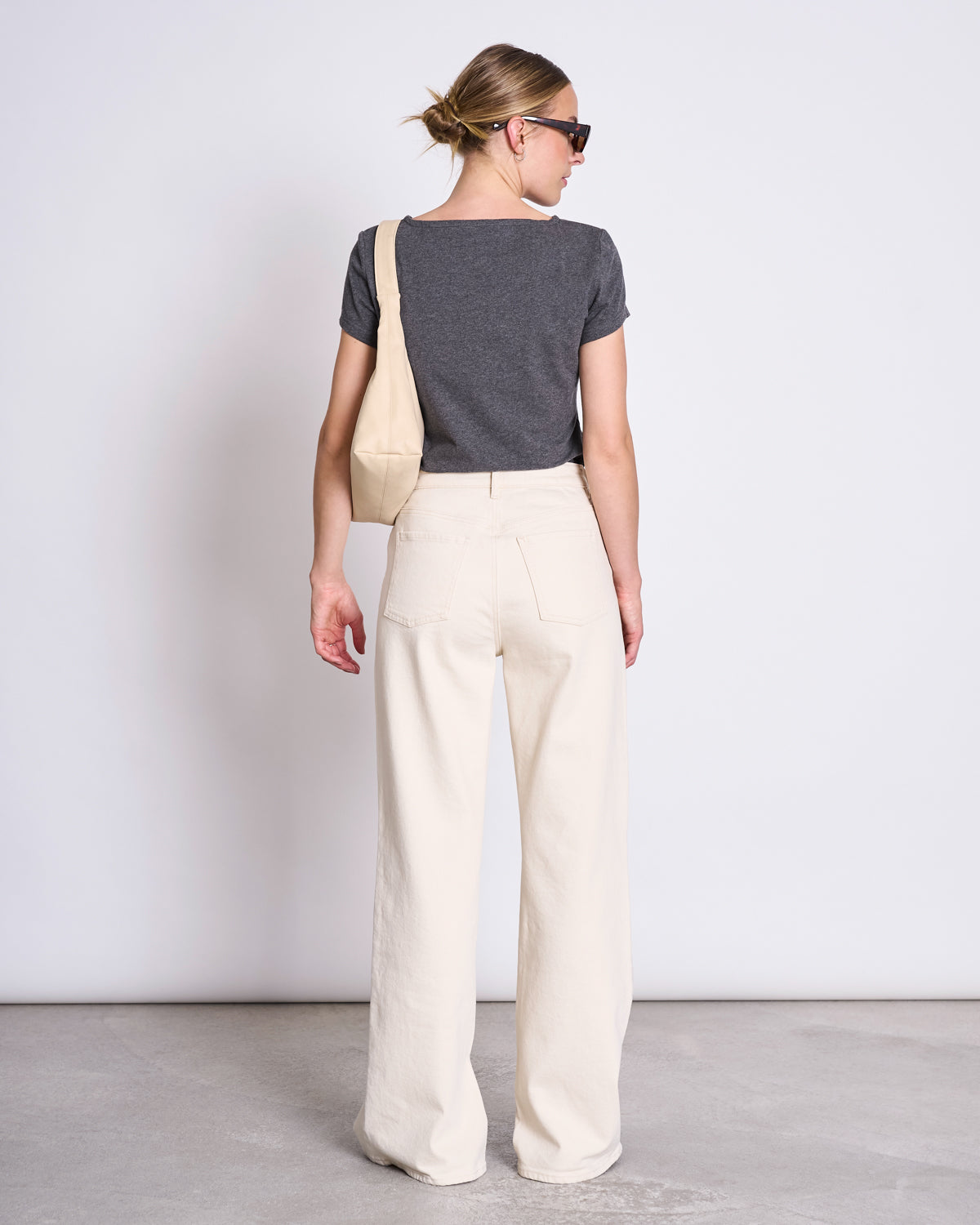 A person faces away from the camera in a jan-n-june T-SHIRT OTTO DARK GREY GOTS, paired with cream wide-leg pants, sunglasses, and a cream shoulder bag against a plain neutral background.