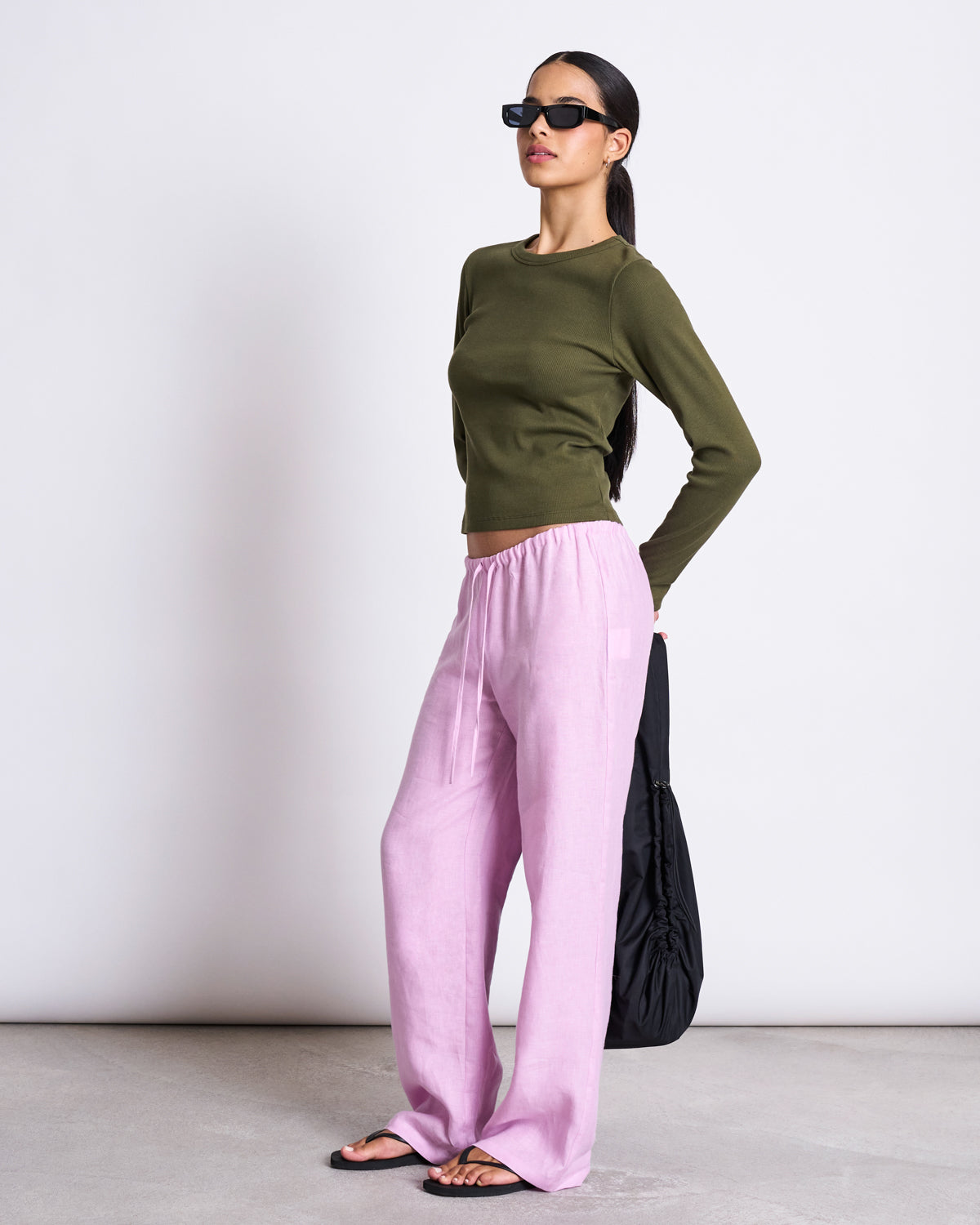 A woman stands against a plain background wearing sunglasses, loose light pink pants, black sandals, holding a black bag behind her back, and the jan-n-june RIB LONGSLEEVE MURI DARK OLIVE GOTS top.