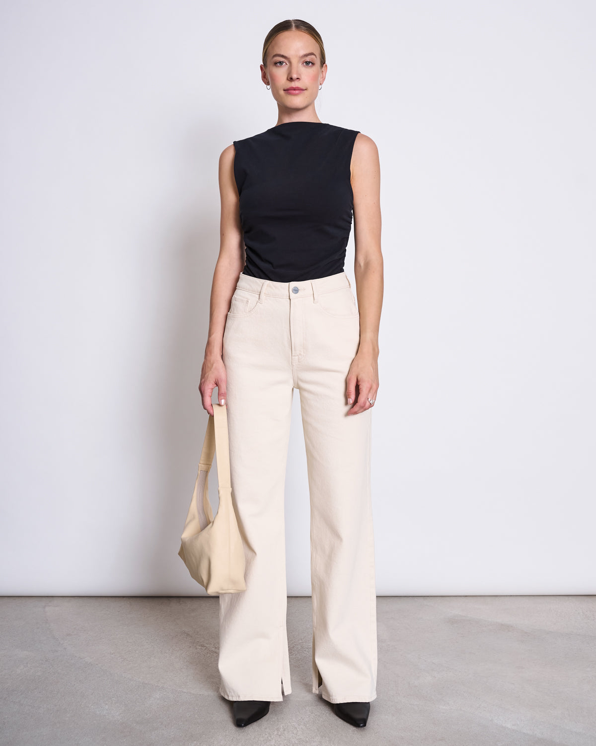 A woman stands against a white wall wearing the jan-n-june RUFFLE TOP RAFO BLACK GOTS with cream wide-leg pants, black shoes, and holding a beige tote bag. Her hair is pulled back as she looks directly at the camera.