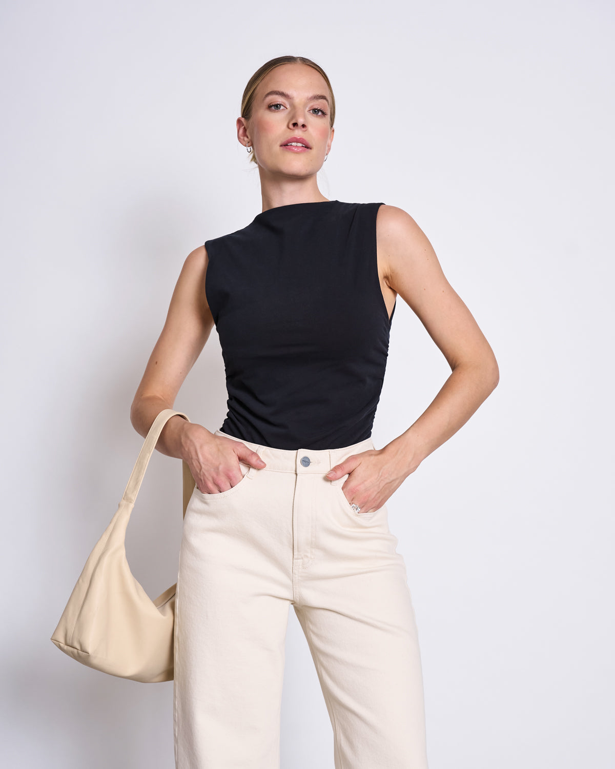 A woman stands against a white background wearing the jan-n-june RUFFLE TOP RAFO BLACK GOTS and high-waisted off-white pants. She carries a beige shoulder bag, hair tied back, and gazes upward with a confident look.
