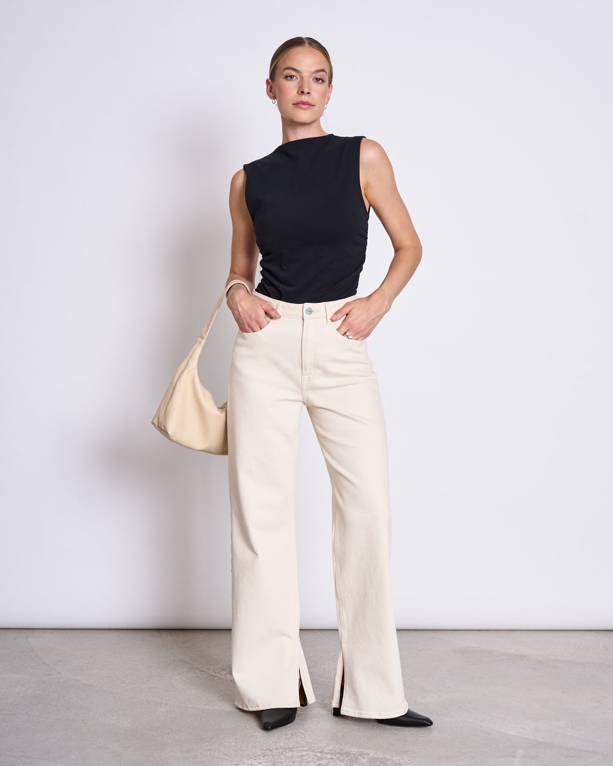A woman poses with one hand on her hip, wearing the jan-n-june RUFFLE TOP RAFO BLACK GOTS, high-waisted cream wide-leg pants with front slits, black shoes, and a beige shoulder bag against a plain white background.