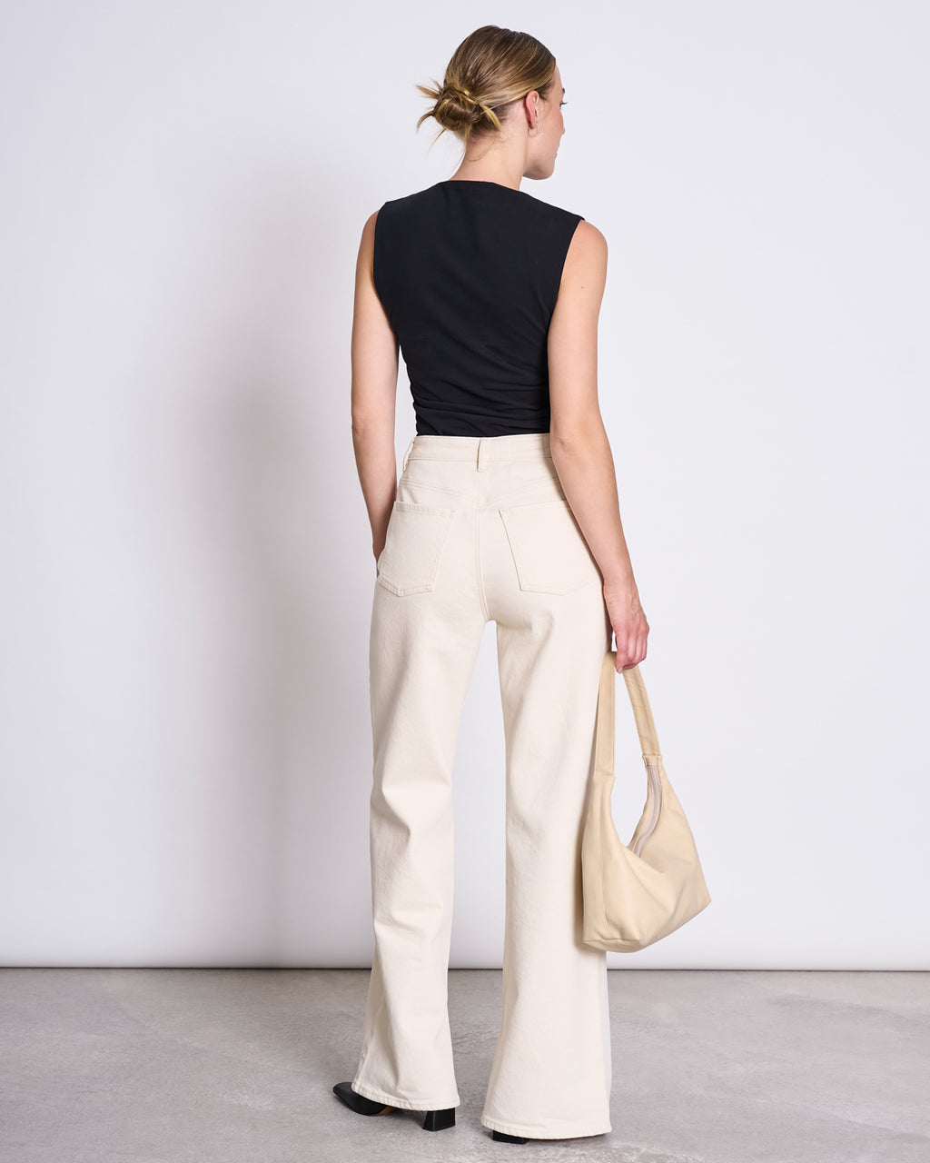 A woman stands facing away, wearing the jan-n-june RUFFLE TOP RAFO BLACK GOTS, high-waisted cream wide-leg pants, black shoes, and holding a beige handbag. Her hair is in a low bun against a plain background.