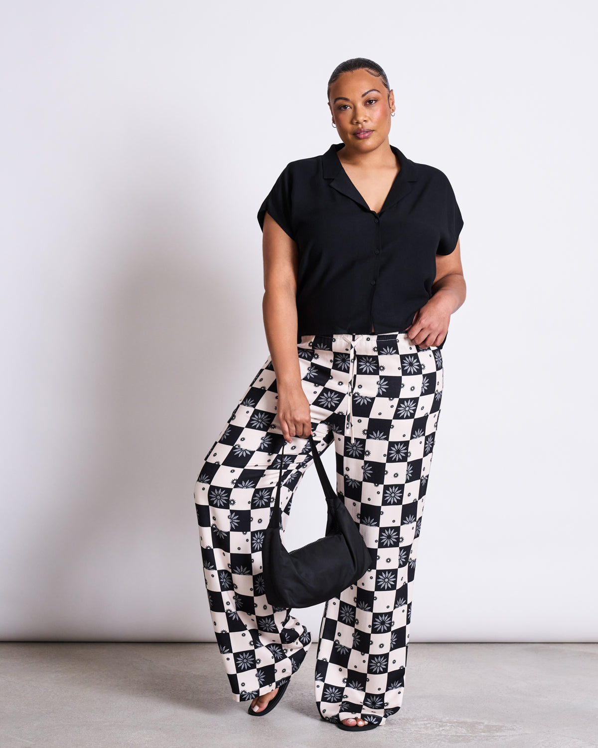 A woman poses against a white wall, wearing jan-n-junes BLOUSE TIERRA BLACK and high-waisted, wide-leg black and white checkered pants with sun and geometric patterns. She holds a black handbag and wears sandals.
