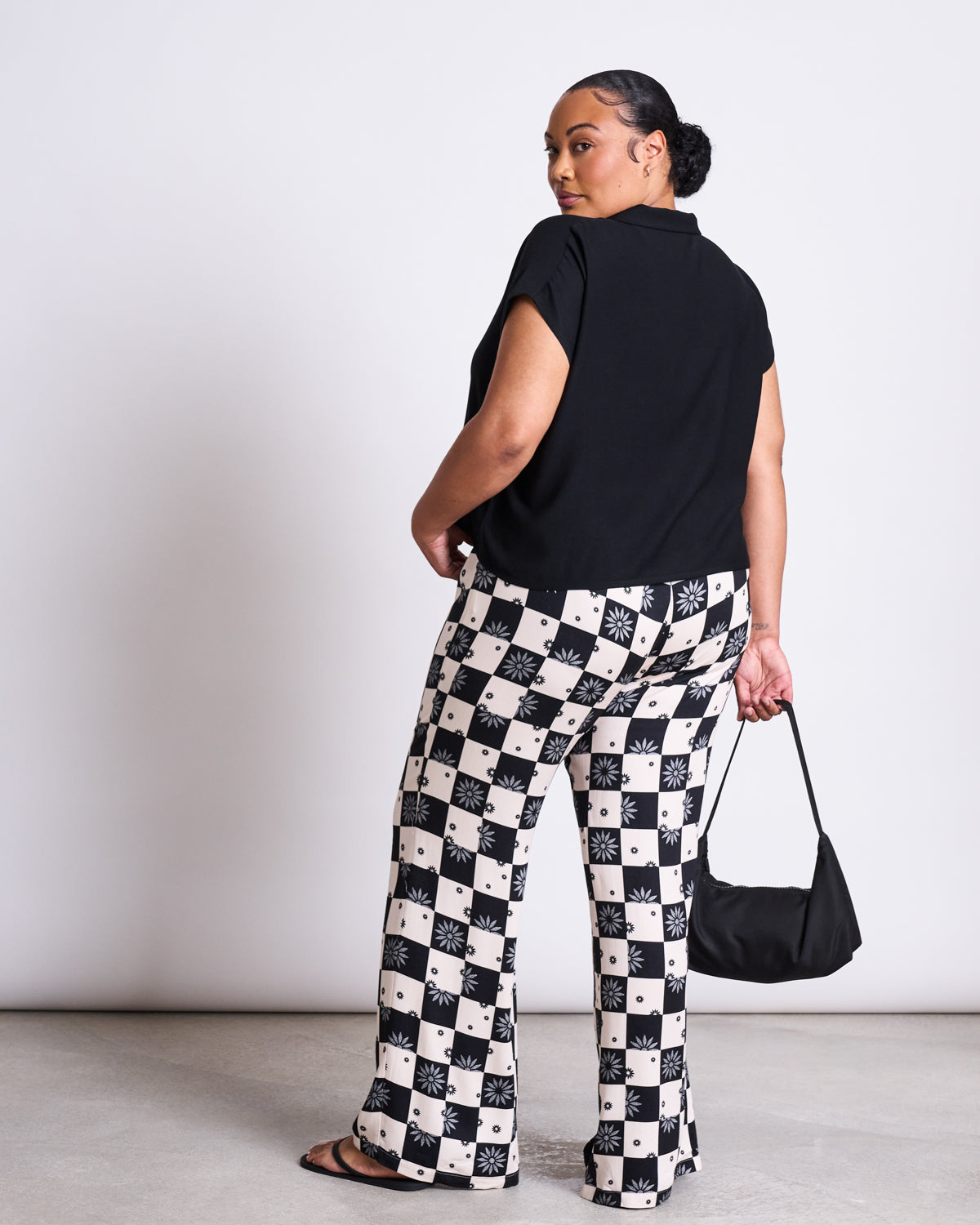 A woman stands slightly turned, wearing the jan-n-june BLOUSE TIERRA BLACK—a black short-sleeved, V-neck blouse—paired with black-and-white checkered floral pants, a small black purse, and black sandals against a plain light background.