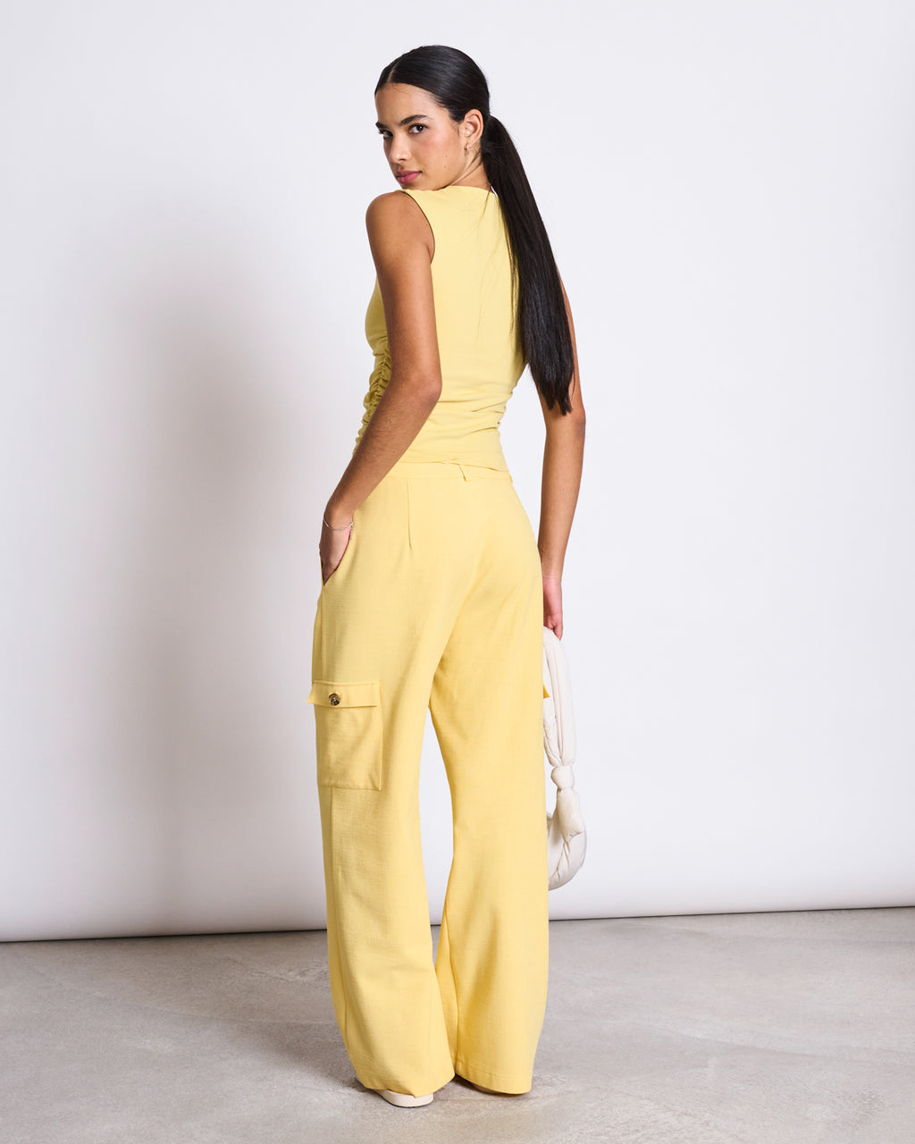 A woman with long dark hair in a ponytail wears a sleeveless yellow top and jan-n-june’s CARGO PANTS FANO BUTTERCUP, wide-leg EcoVero pants with a side pocket, standing sideways and looking over her shoulder against a plain background.