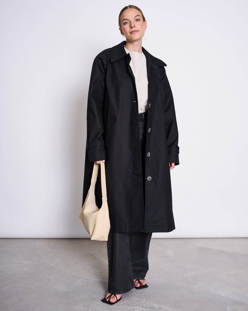 A woman stands against a plain background wearing the jan-n-june COAT TAGUS BLACK, an oversized long black transitional coat, over a white top and dark pants. She holds a light beige handbag and wears black open-toe shoes.