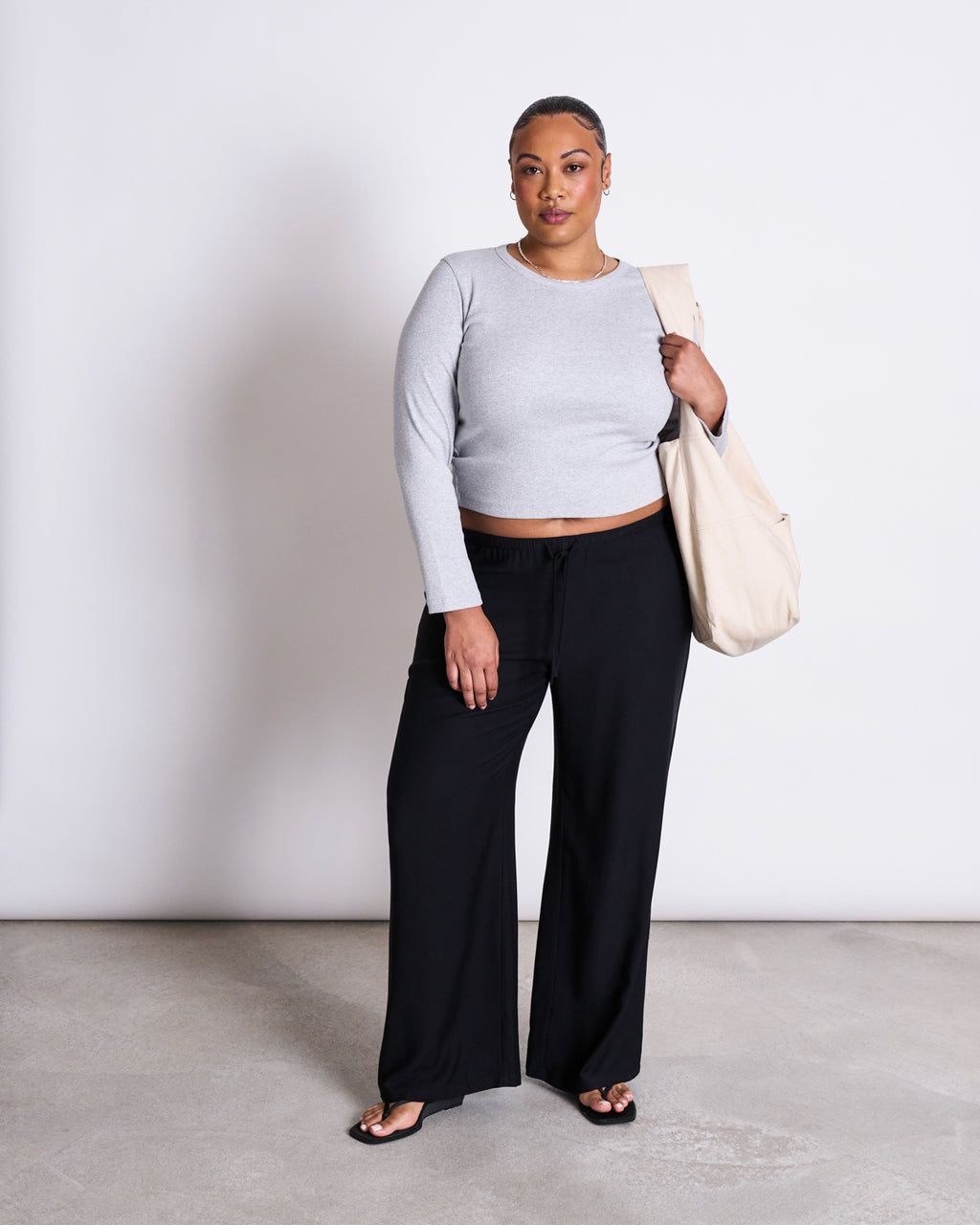 A woman stands against a plain white background, wearing MID RISE LOOSE PANTS STRATH BLACK by jan-n-june, a light gray MicroModal long-sleeve top, black sandals, and carrying a large beige tote bag over her shoulder.