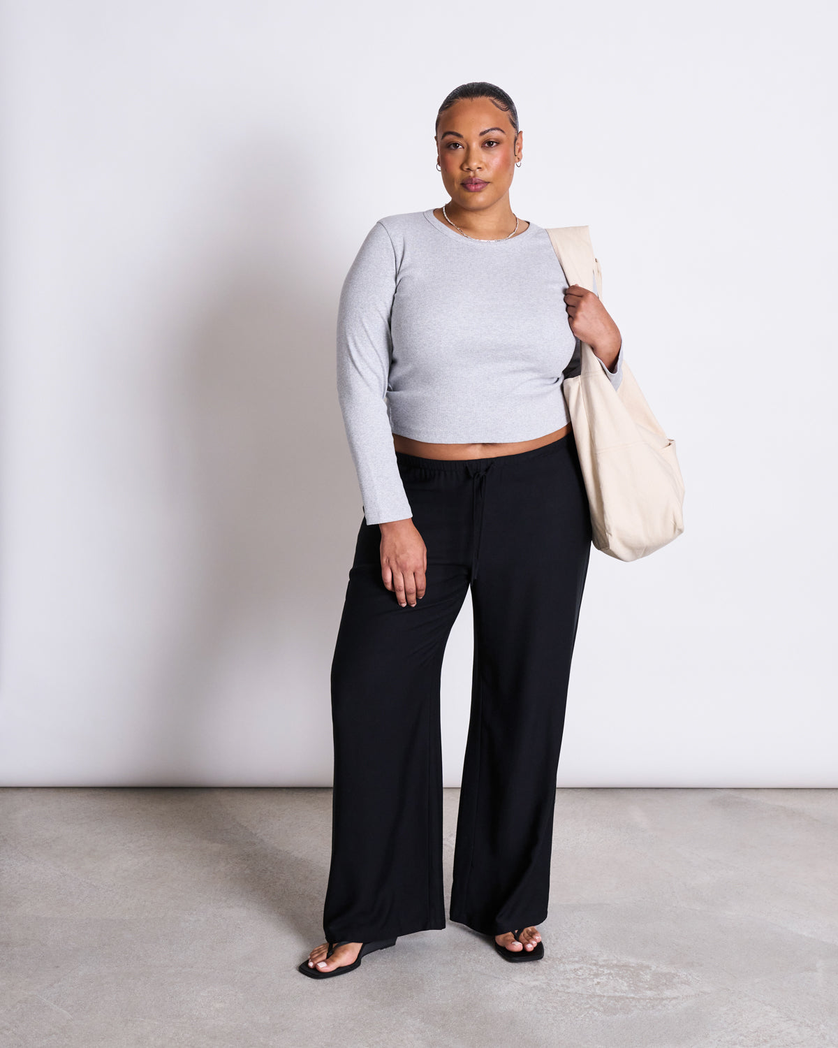 A woman stands against a plain white background, wearing MID RISE LOOSE PANTS STRATH BLACK by jan-n-june, a light gray MicroModal long-sleeve top, black sandals, and carrying a large beige tote bag over her shoulder.