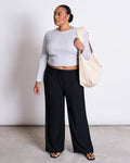 A woman stands against a plain background wearing the jan-n-june RIB LONGSLEEVE MURI LIGHT GREY GOTS crop top, loose black pants, and sandals. She carries a large beige tote bag over her shoulder and looks to the side.