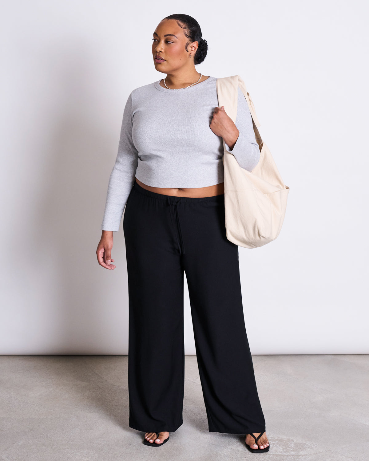 A woman stands against a plain background wearing jan-n-junes MID RISE LOOSE PANTS STRATH BLACK, a long-sleeve gray crop top, sandals, and a cream tote bag on her shoulder. She looks slightly to the side.