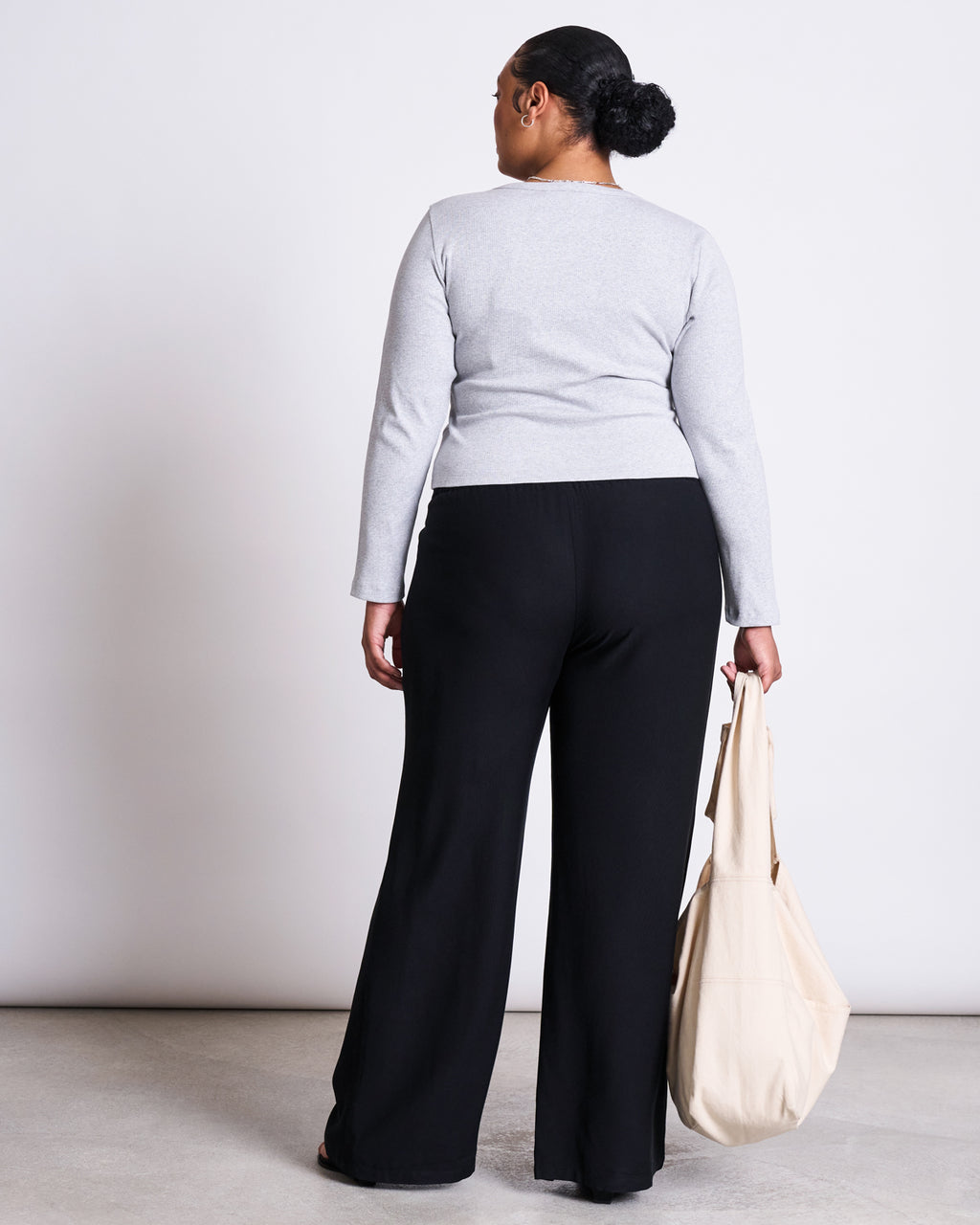 A woman with her back to the camera wears a light gray long-sleeve top and jan-n-june MID RISE LOOSE PANTS STRATH BLACK, holding a beige tote bag in her left hand. She stands on a light floor against a plain white background.