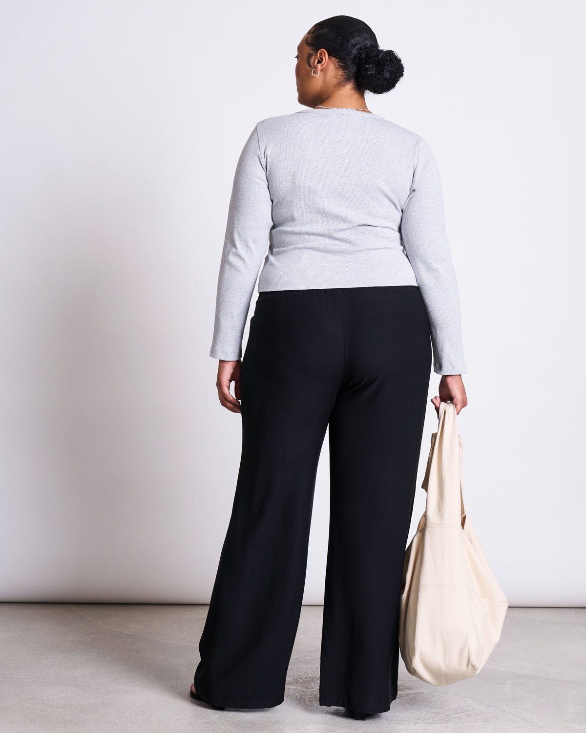 A woman with her back to the camera wears a light gray long-sleeve top and jan-n-june MID RISE LOOSE PANTS STRATH BLACK, holding a beige tote bag in her left hand. She stands on a light floor against a plain white background.