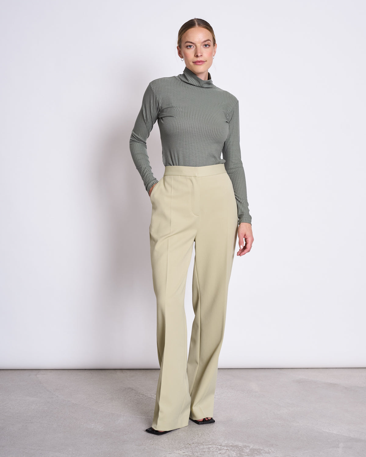 A woman poses against a white background wearing the jan-n-june LYOCELL RIB TURTLENECK MIO SEA SPRAY in sage green and high-waisted, wide-leg beige trousers, with her hands in her pockets.