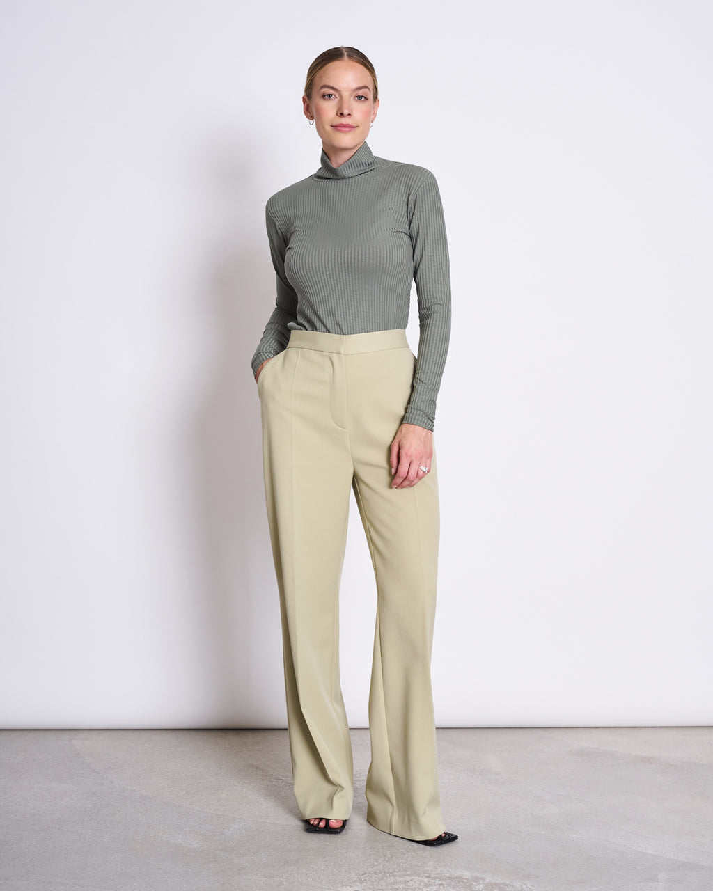A woman stands against a white background wearing the jan-n-june LYOCELL RIB TURTLENECK MIO SEA SPRAY in sage green, paired with high-waisted beige wide-leg trousers and black open-toe shoes, with her hands in her pockets.