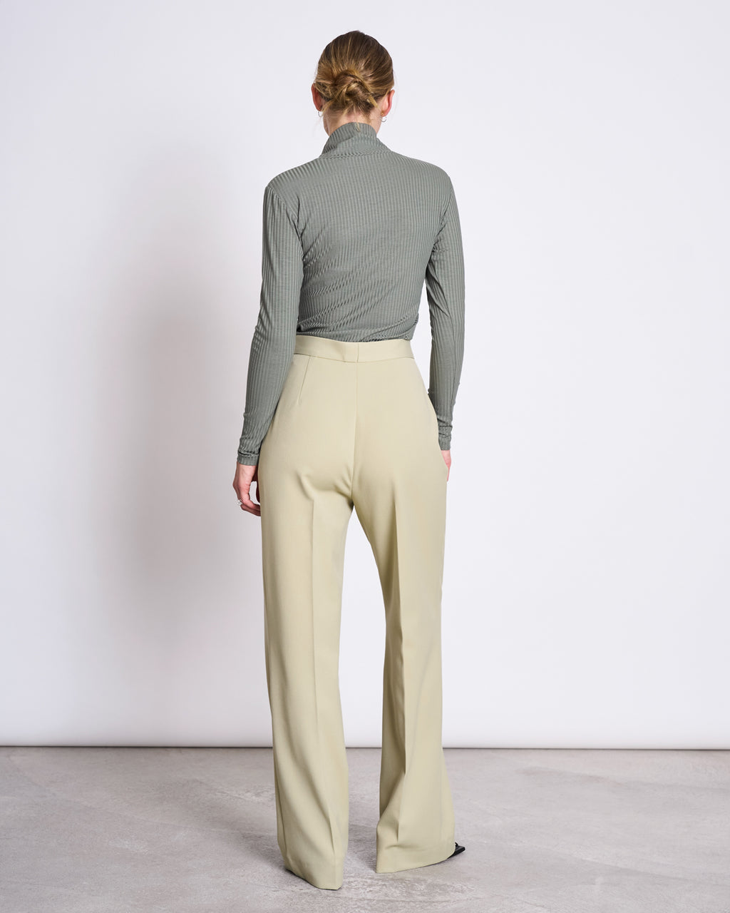 A woman faces away from the camera, wearing the jan-n-june LYOCELL RIB TURTLENECK MIO SEA SPRAY top with high-waisted, wide-leg beige trousers, standing on a light floor against a plain white wall.