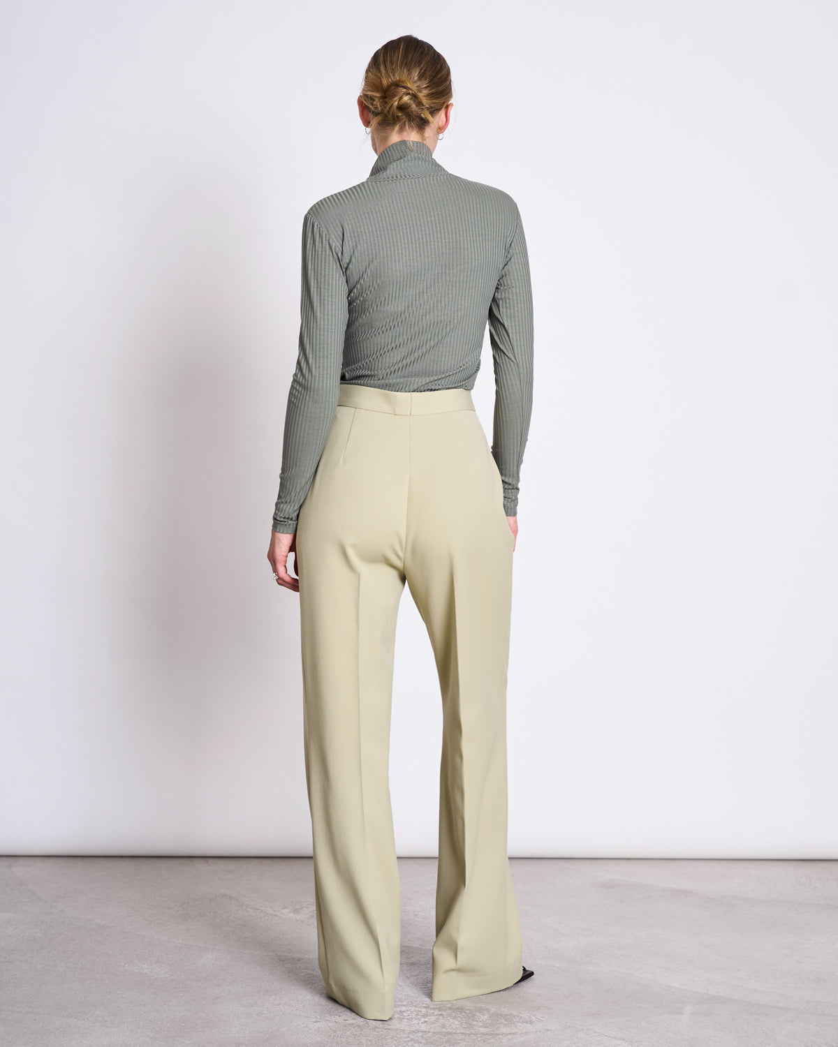 A woman faces away from the camera, wearing the jan-n-june LYOCELL RIB TURTLENECK MIO SEA SPRAY top with high-waisted, wide-leg beige trousers, standing on a light floor against a plain white wall.