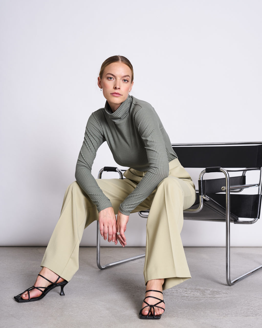 A woman wearing the jan-n-june LYOCELL RIB TURTLENECK MIO SEA SPRAY and beige pants sits on a modern black and chrome chair, one arm on her knee, the other hanging down, looking directly at the camera.