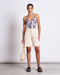 A woman stands against a plain background wearing the jan-n-june TOP MATILDA MELTED PRINT GOTS, paired with high-waisted beige frayed hem shorts, black platform sandals, and holding a light-colored handbag.