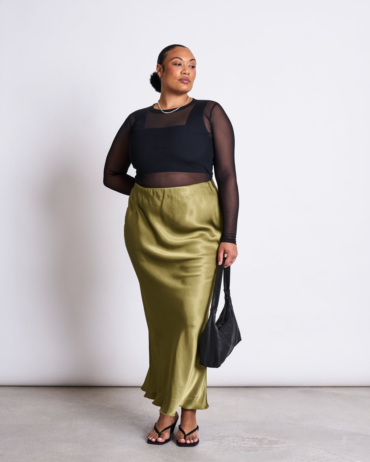 A woman poses confidently against a plain background wearing the jan-n-june MAXI SKIRT SERIN WOODY OLIVE, paired with a sheer black long-sleeve top over a black bra, black sandals, and holding a black handbag.