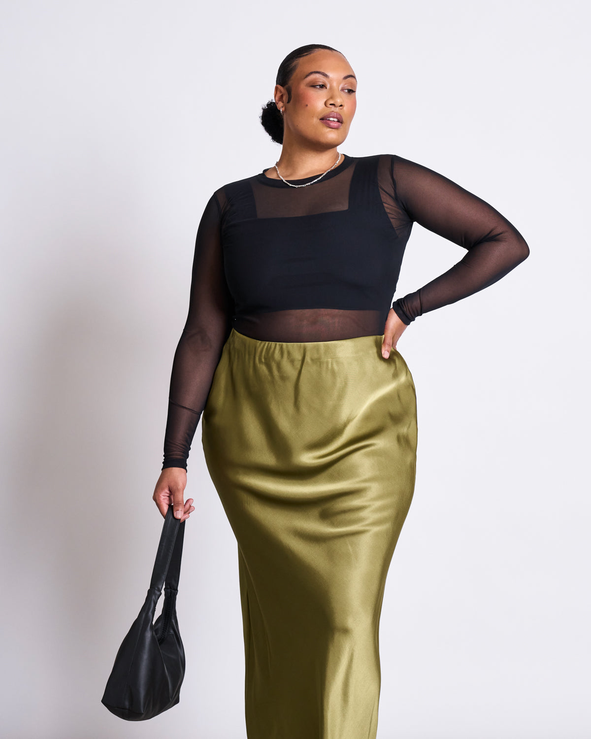 A woman poses confidently in a jan-n-june MAXI SKIRT SERIN WOODY OLIVE, paired with a sheer black long-sleeve top and black handbag. She accessorizes with a necklace and sleek hair—an ideal look for summer weddings.