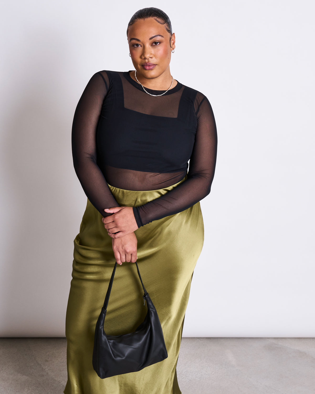 A woman poses against a plain background, wearing the jan-n-june MAXI SKIRT SERIN in woody olive with a sheer black long-sleeve top and a small black handbag—an elegant look ideal for summer weddings.