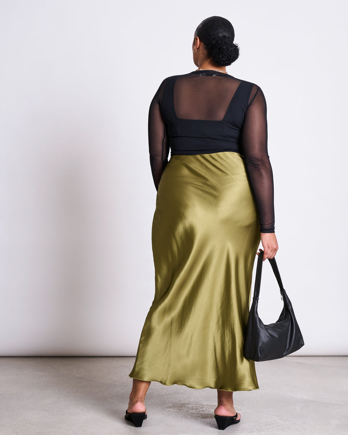 A woman in a black sheer top and the jan-n-june MAXI SKIRT SERIN WOODY OLIVE stands with her back turned. She holds a black handbag, wears black sandals, her hair in a sleek bun. Set against a plain background—ideal for summer weddings.