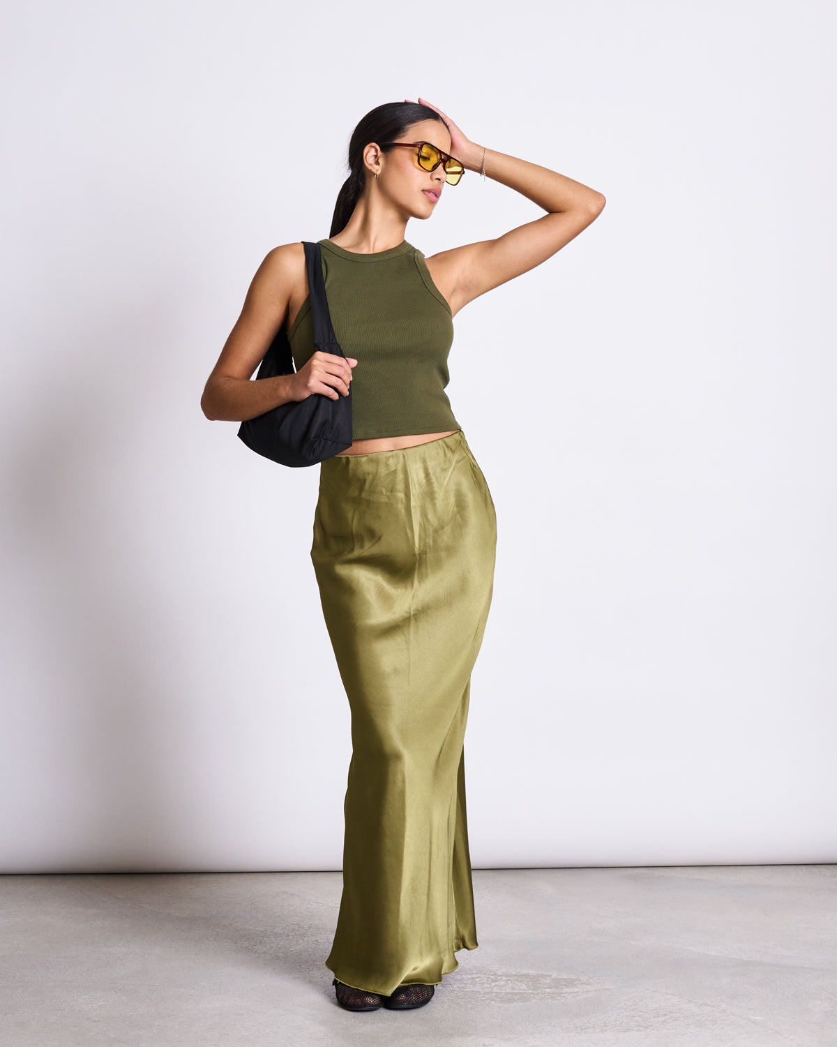 A woman wearing jan-n-junes MAXI SKIRT SERIN WOODY OLIVE, paired with a sleeveless crop top, black bag, and yellow-tinted sunglasses, poses in a minimalist studio—an ideal look for summer weddings.