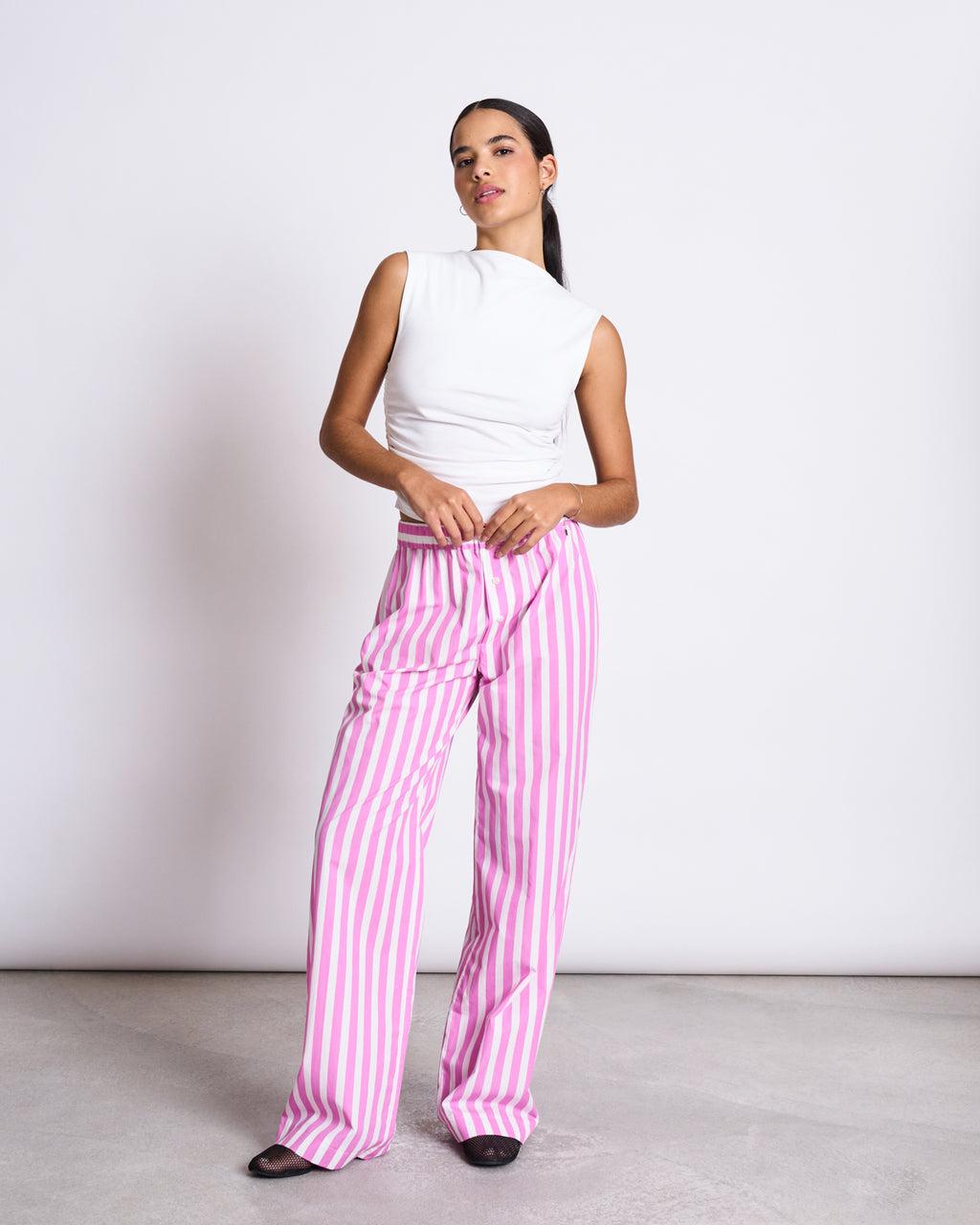 A woman stands confidently with hands at her waist, wearing jan-n-june’s COTTON PANTS SOBY WHITE HOT PINK STRIPED GOTS—high-waisted, wide-leg organic cotton pants with vertical pink and white stripes.