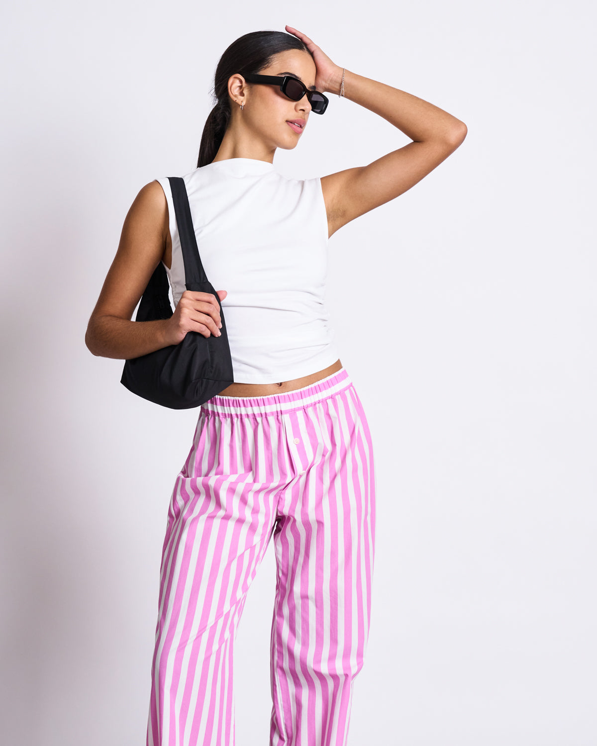 A woman models jan-n-junes COTTON PANTS SOBY WHITE HOT PINK STRIPED GOTS with a sleeveless white top, sunglasses, and a black bag, posing with one hand on her head against a plain white background.
